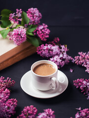 A white coffee cup, blooming branches of purple lilac on a concrete black table with old books