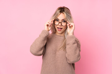 Teenager blonde girl wearing a sweater over isolated pink background with glasses and surprised