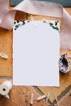 A blank white letterhead lies on a stone tile framed by a ribbon, a shell and a skein of thread