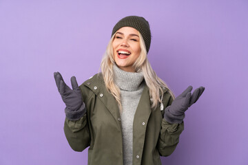 Teenager blonde girl with winter hat over isolated purple background smiling a lot