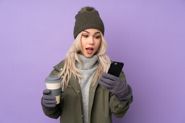 Teenager blonde girl with winter hat over isolated purple background holding coffee to take away and a mobile