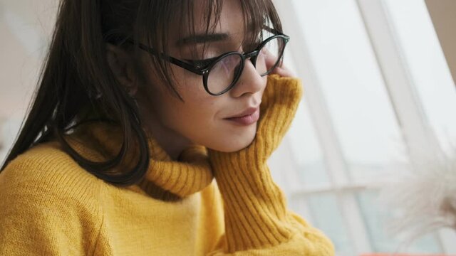 The close-up shot of the face of a smiling woman with glasses and a yellow sweater with her hand on her face while sitting in the studio
