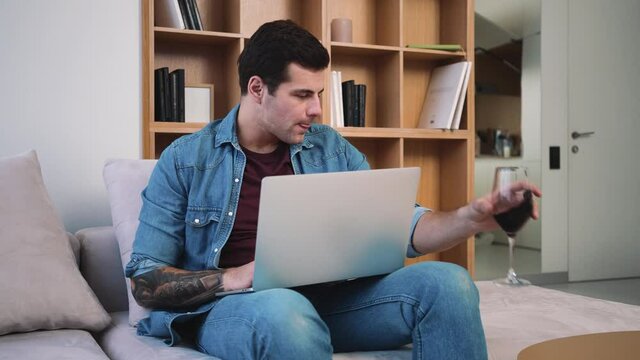 A pleased man is using his laptop computer while drinking a glass of wine sitting on the sofa inside the apartments
