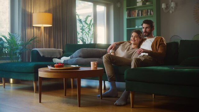 Couple Watches TV while Sitting on a Couch in the Living Room. Girlfriend and Boyfriend lying embracing, switch Channels with Remote Control. Spending Weekend Together at Home with Stylish Interior.