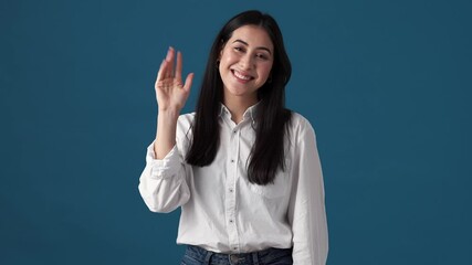 A smiling korean woman is waving hand showing goodbye gesture standing isolated over blue wall in studio
