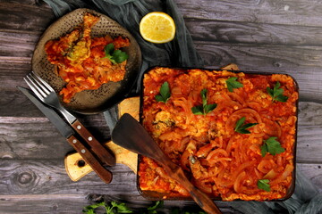 Dough baked herring fish with tomato sauce and onion in a glass bowl. Fish, baked with vegetables in tomato sauce. Dark wooden background. Top view. 