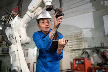 Fototapeta premium Professional woman check the robotic arm machine in the factory. Worker wearing safety helmet, glasses and uniform. Preventive maintenance to prevent breakdown. Service and repair concept