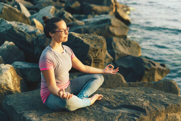 Young woman sitting on a stone pier by the sea meditates in the lotus position. Brunette girl doing...