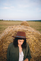 Stylish woman in hat standing at hay bale in summer evening in field. Tranquility. Rural slow life