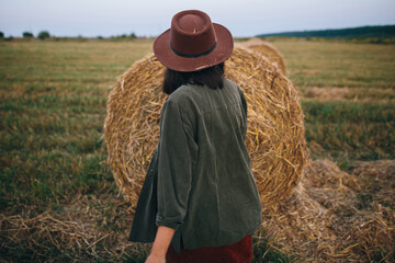 Naklejka na ściany i meble Beautiful stylish woman in hat walking at hay bales in summer evening field. Vacation in countryside