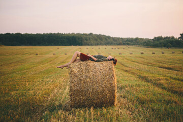 Beautiful carefree woman lying on haystack enjoying evening in summer field. Atmospheric tranquility