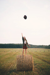 Fototapeta na wymiar Beautiful carefree woman throw hat up in the sky standing on haystack in summer field. Happiness