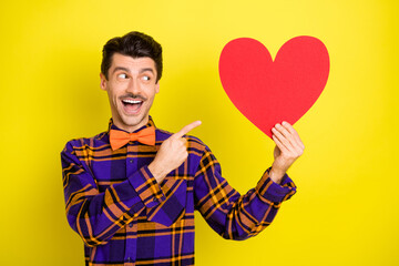 Photo of guy hold red paper heart card direct finger wear bow tie plaid shirt isolated yellow color background