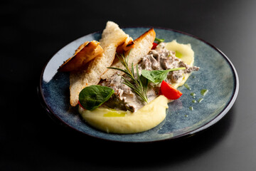 Meat in a creamy sauce with mashed potatoes and bread toast, painted with spinach leaves and cherry tomatoes, on a gray plate on a black background.
