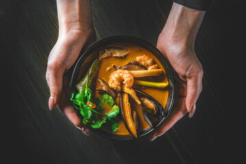 pan asian food. woman hands holding Tom Yam soup in black bowl on wooden table background