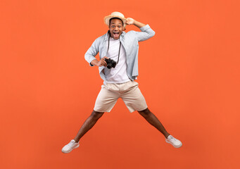 Fototapeta na wymiar Full length of excited black man in summer outfit holding professional camera and jumping on orange studio background