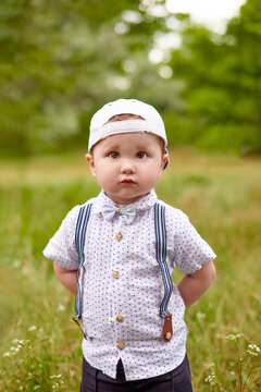 little serious boy in suspenders and a cap hides his hands behind back in park