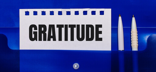 On a blue background, white pens and a sheet of paper with the text GRATITUDE