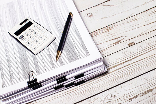 A White Calculator And A Pen Lie On A Stack Of Documents On A White Wooden Table. Business Concept