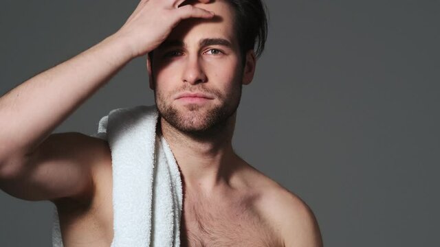 A good-looking man is posing to the camera with a towel on his shoulders standing isolated over grey wall in the studio