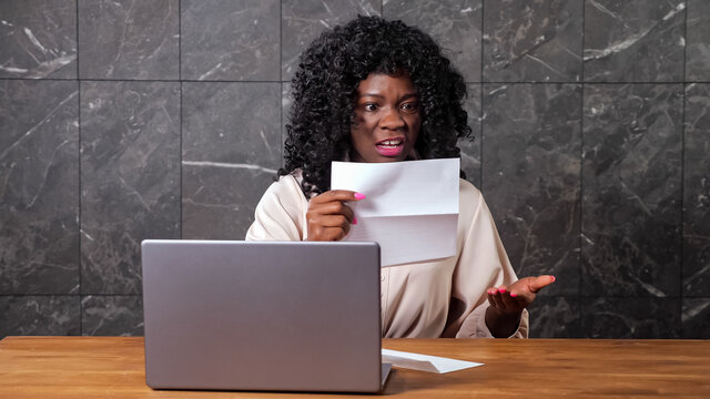 Stressed Afro-american businesswoman opens envelope with bank letter and enters code on grey laptop keyboard on wooden table against marble office wall