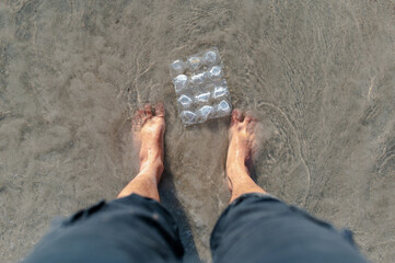 Top view man's feet on the beach surrounded by plastic garbage.