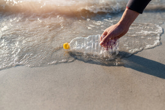 Unrecognizable man cleaning the beach from waste. Close-up hand collecting an empty plastic bottle from the shoreline.