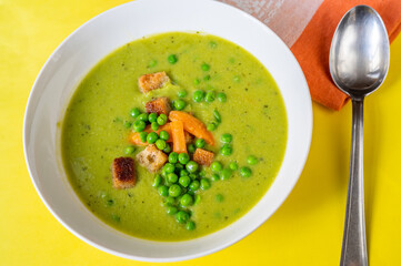 Plate with soup from young pea, spoon and towel on yellow background.