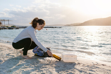 young woman on the beach shore during sunset recollecting waste and putting it into a bag.