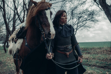Woman in image of medieval warrior stands near horse among forest.