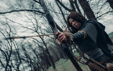 Woman in image of medieval warrior archer shoots a bow in forest.