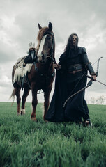 Woman in image of medieval warrior stands with bow near horse.