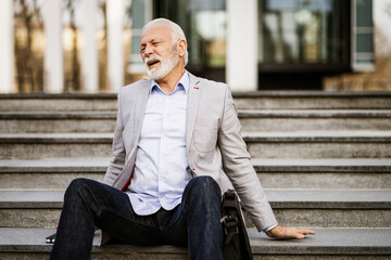 Obraz premium Senior businessman is tired of work. He is sitting on stairs in front of company building.