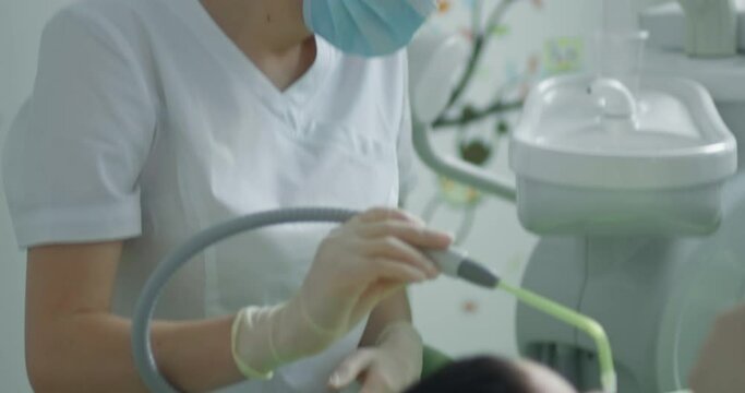 A female medical worker is taking a saliva ejector and putting it into the mouth of the little boy who is laying in the dental chair while a dentist is sitting near the boy and holding a dental tool.