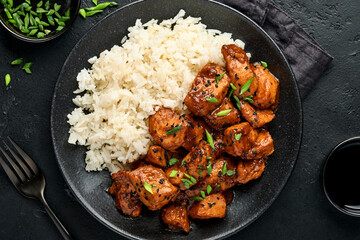 Spicy teriyaki chicken fillet pieces with rice, green onions and black sesame seeds on black plate on a dark slate, stone or concrete background. Top view with copy space.