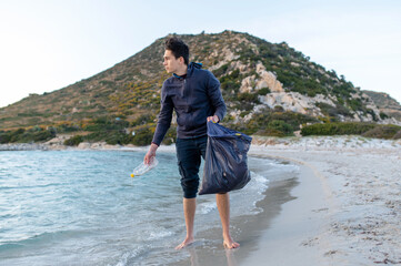 Young man standing on the beach shore recollecting plastic garbage into a bag. Volunteer work and environmental conservation