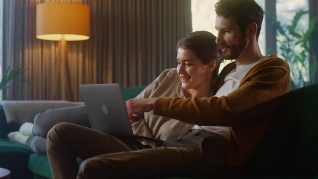 Young Couple Use Laptop Computer, while Sitting on a Couch in the Cozy Stylish Apartment. Boyfriend and Girlfriend Shopping on Internet, Using Social Media, Watching Funny Videos and Streaming Service