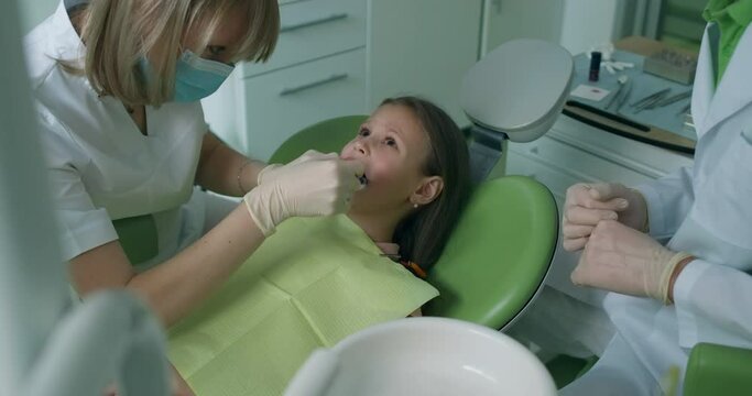 A little girl is laying in the dental chair while the female dentist is doing an injection and after it is giving a syringe to the male assistant.