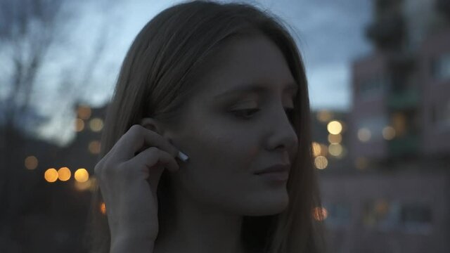 Close up, young pretty woman placing bluetooth earphone in ear in city twilight exterior, slow motion