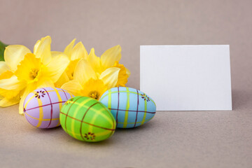 There are colorful Easter eggs next to a bouquet of yellow daffodils, and next to them is a white card with a place for the inscription. close-up. Place for your text. Postcard.