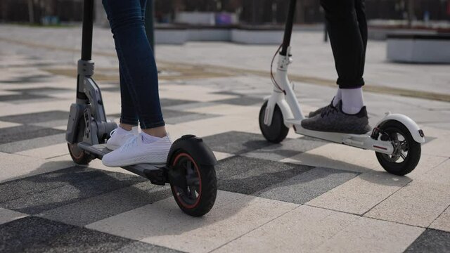 A man and a woman ride electric scooters. In the frame, the legs and footboards of scooters.