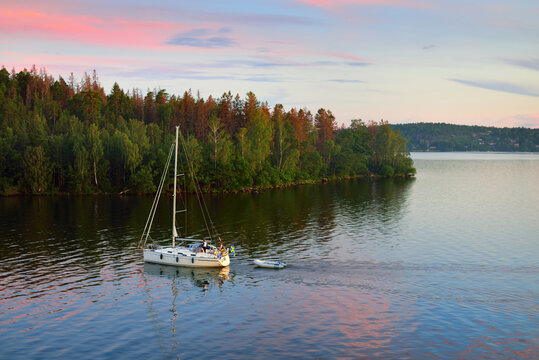 Sloop rigged yacht with inflatable boat sailing at sunset. Panoramic aerial view of the forest lakeshore. Moonrise, glowing clouds. Mälaren lake, Sweden. Summer vacations, nature, sailing, cruise