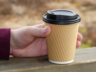 Cardboard cup for hot drinks in the left hand of a young man on a blurred background