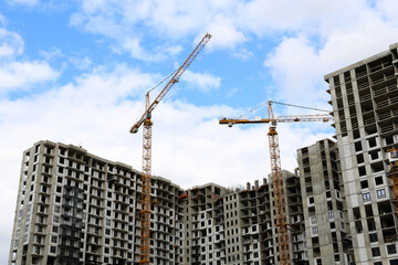 Construction cranes above unfinished residential buildings on blue sky and white clouds background. Housing construction, apartment block in city