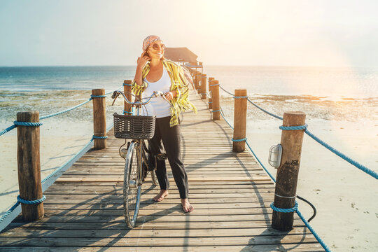 Portrait of a smiling woman dressed in light summer clothes, sunglasses with bicycle on the wooden sea pier on the sandy Zanzibar beach.Careless vacation in the tropical countries concept image