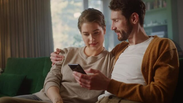 Couple Use Smartphone Device, while Sitting on a Couch in the Cozy Apartment. Boyfriend and Girlfriend Talk, do e-Shopping on Internet, Watching Funny Videos, Use Social Media, Streaming Service.