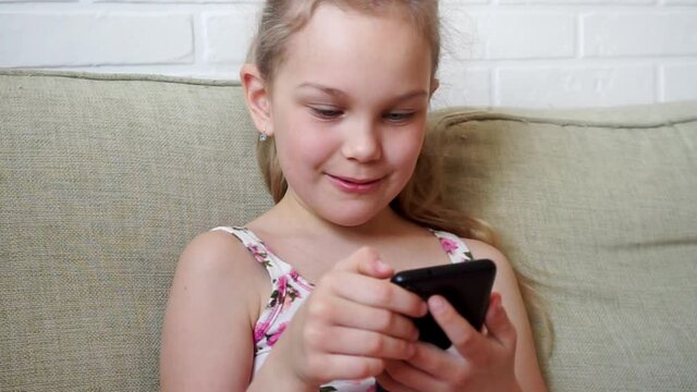 Girl playing mobile game on smartphone on sofa. Preschooler playing mobile phone. Kid using phone for gaming. Child playing video game at home
