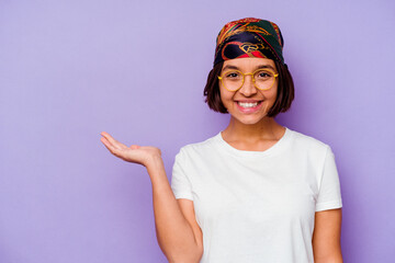 Young mixed race woman wearing a bandana isolated on purple background showing a copy space on a palm and holding another hand on waist.