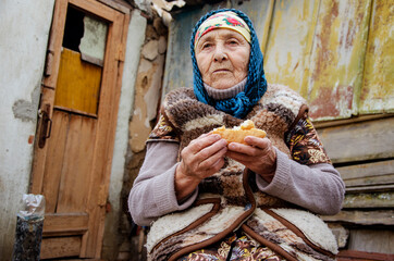 Grandmother eats bread against the backdrop of slums. The old grandmother is eating a piece of bread. Hunger and poverty. Slums. Poverty. Unprotected beliefs of the population. Need food. Poor areas.