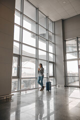 Fototapeta na wymiar Caucasian woman is standing alone near airport terminal panoramic windows with luggage
